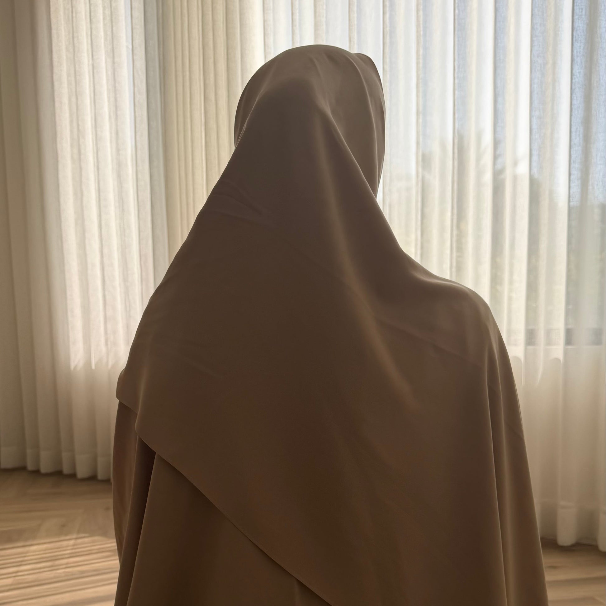 Person wearing a brown hijab standing in front of a window with light curtains.